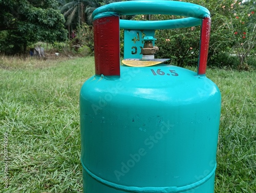A green 16.5 kg LPG gas cylinder placed upright on a grassy field outdoors. The steel liquefied petroleum gas cylinder features a protective ring around the valve . in malaysia