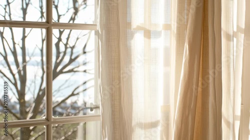Slow motion tracking shot of linen curtains swaying and billowing at sunlit window overlooking seaside view from interior room