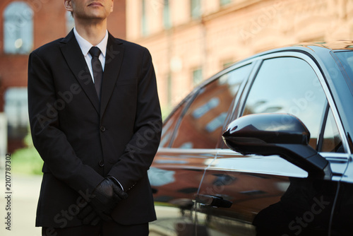 Foto Caucasian young adult chauffeur standing beside luxury sedan, waiting for passenger pickup in urban setting