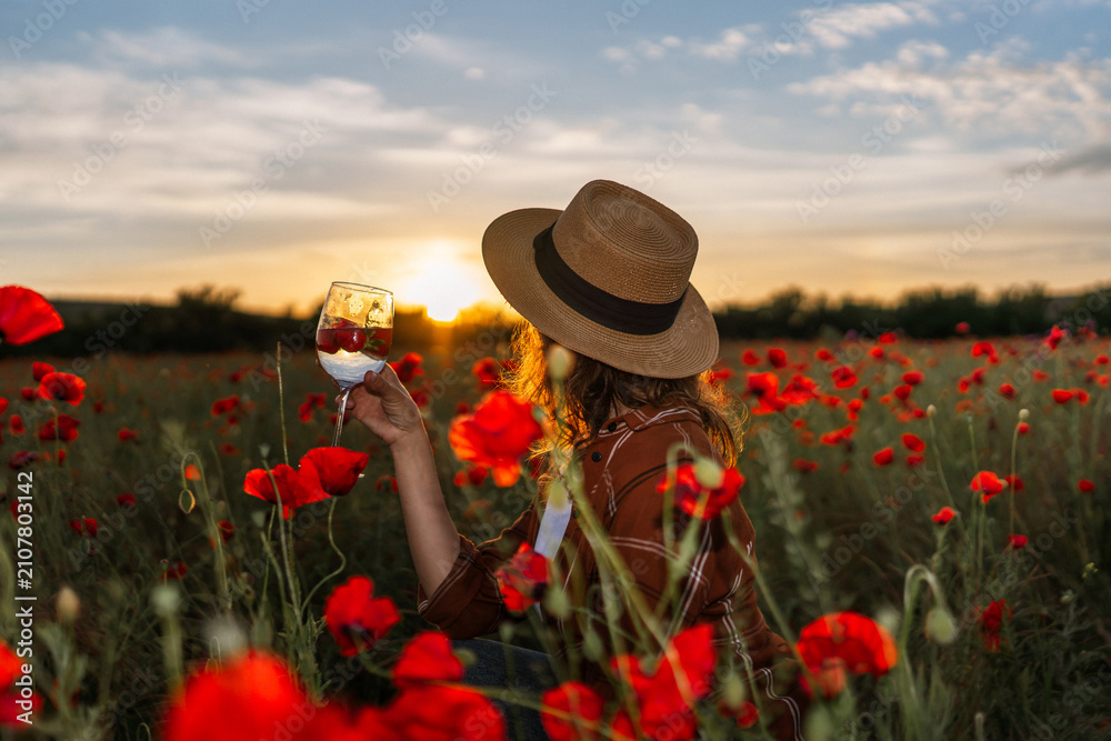 Obraz premium Golden hour sunset features woman wearing straw hat holding wine glass in field of Papaver rhoeas