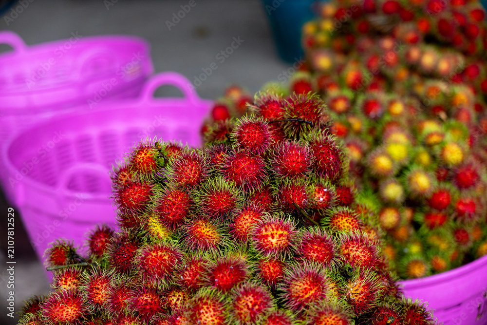 Naklejka premium Rambutan fruit pile with vibrant red spiky skins and green hair displayed in purple plastic baskets at market, colorful tropical produce and fresh harvest evoke lively atmosphere