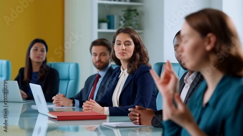 Female professional in blue blouse gestures while speaking during a business meeting with colleagues seated at a conference table in a modern office setting