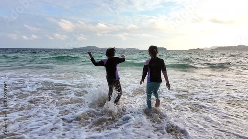 Two Asian girls playing and splashing water in ocean waves on sandy beach during summer vacation