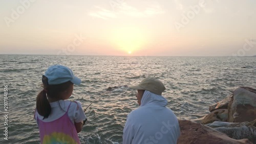 Mother and daughter fishing together with fishing rod on rocky seashore at evening