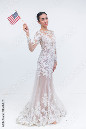 Fotografie A young Southeast Asian bride in a detailed lace wedding gown holds and waves a small American flag while standing in a graceful contrapposto pose