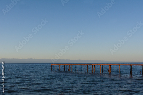 Bild auf Leinwand Long wooden pier extending into calm ocean waters under a clear blue sky with distant mountains on the horizon during a peaceful sunny day