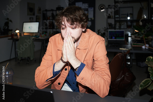 Young adult Caucasian man covering mouth while staring at laptop in office, showing stress after work issue. Useful for burnout, deadline pressure, remote work crisis marketing