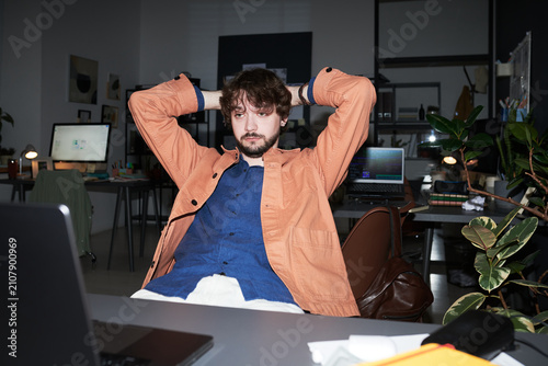 Young adult Caucasian man leaning back with hands behind head, looking at laptop in office, reflecting on workload. Useful for remote work, burnout, productivity themes