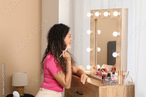 Beautiful young happy African-American woman with brush doing makeup near mir...