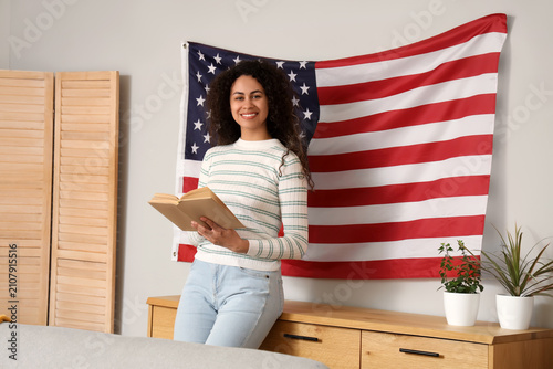 Young African-American woman reading book near hanging USA flag on wall at home