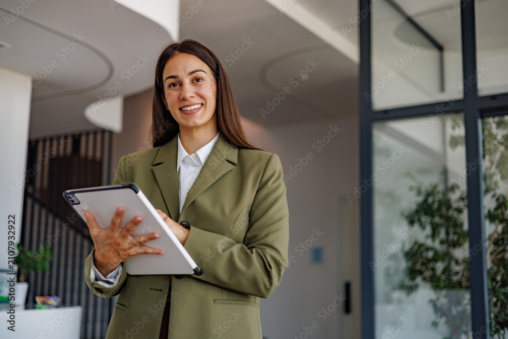 Obraz premium Businesswoman standing in modern office holding a tablet, smiling and looking at camera while working