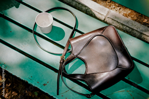 A leather handbag and a disposable cup rest on a green park bench