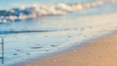 Close up of soft ocean waves gently washing onto sandy beach shore with sunlight reflecting on water