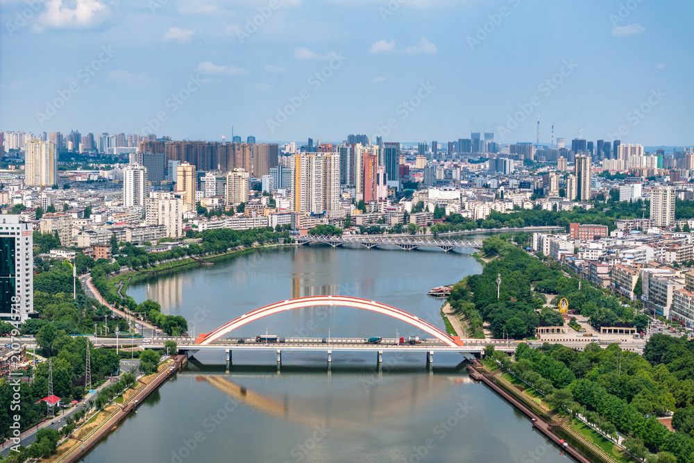 Obraz premium Aerial view of a modern city with an arch bridge crossing a calm river, surrounded by buildings and green spaces under clear sky.