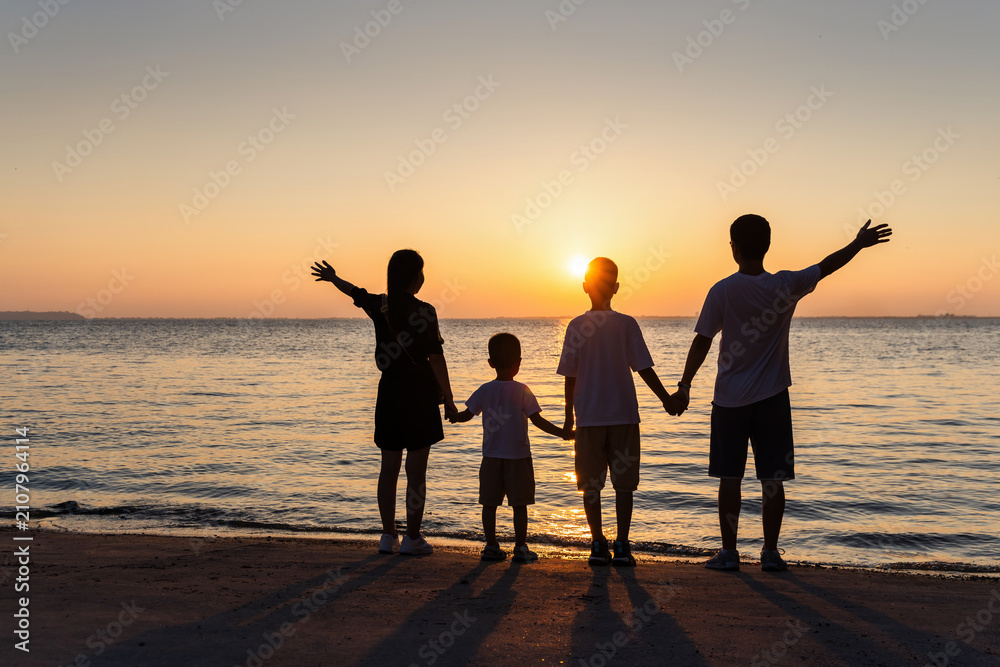 Fototapeta premium A family group of five stands on a beach at sunset, holding hands, with arms raised against the glowing sky and calm sea.