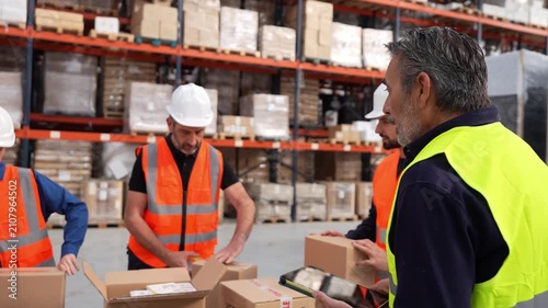 Group of warehouse employees packing fragile items into cardboard boxes while a manager oversees the shipping process using a tablet