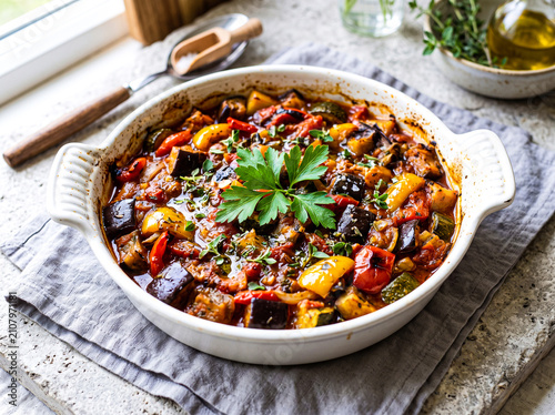 Delicious homemade ratatouille in a white ceramic dish garnished with fresh herbs and placed on a rustic kitchen countertop with natural light streaming through a nearby window