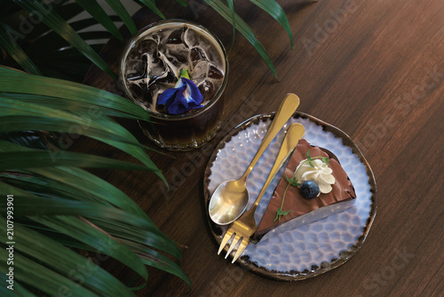 Fotografía A delicious slice of chocolate cake with whipped cream, blueberry, and an iced coffee with a butterfly pea flower on a wooden table with lush green leaves