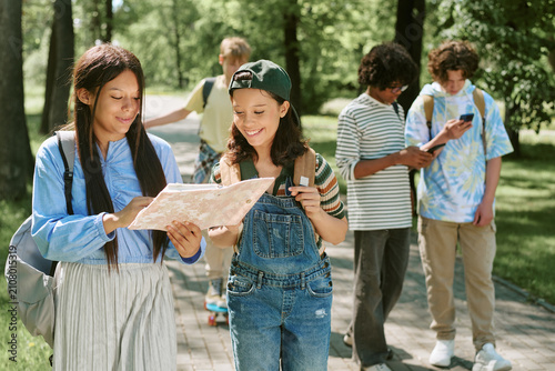 фотографія Diverse teen students studying outdoors, reading notes in copybook and checking smartphone while walking in park, representing teamwork and learning