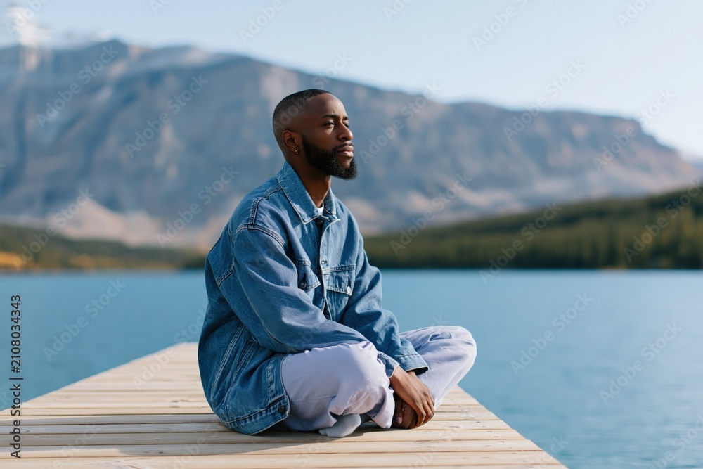 Fototapeta premium Side profile male portrait seated on dock by alpine lake in denim jacket for meditation background and wellness banner
