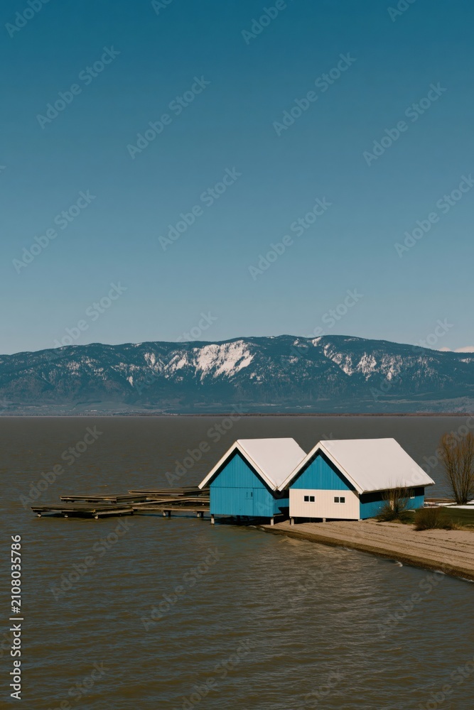 Fototapeta premium Blue boathouse duo on a calm lake with snow-capped mountain background for lakeside wallpaper and banner design