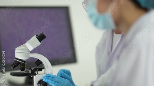 A scientist concentrates at a microscope beside a monitor showing magnified purple cell tissue in a pathology laboratory.