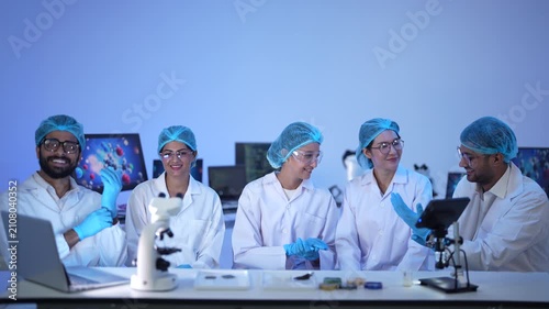 A diverse team of five scientists sit together at a bench with microscopes and colourful samples, smiling to camera under blue light.