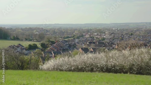 Rows of brick houses sit among greenery with distant hills and open fields beyond. Clear skies highlight the blend of suburban living and rural landscape.