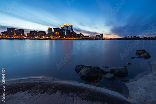 Kalasatama skyline across the Baltic Sea bay at twilight, long exposure photograph of modern skyscrapers, rocky shoreline, and illuminated city lights in Helsinki, Finland.