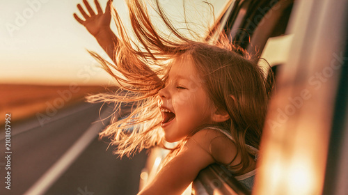 A happy girl leans out of the window of a moving car at sunset, her hair blowing in the wind, a sense of freedom, childhood, and a family trip.