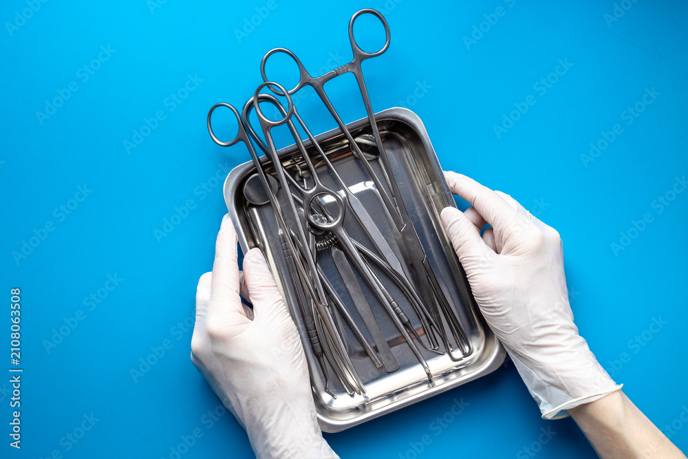 Obraz premium Hand in white glove is holding sterile surgical instruments and tools - scalpels, scissors and others - in a stainless tray, top view