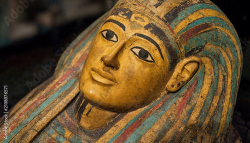 Close-up of an ancient Egyptian sarcophagus featuring a painted yellow face, kohl-rimmed eyes, weathered wood, and intricate green and red headpiece details.