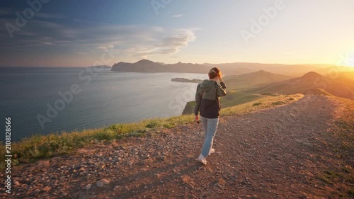 Young woman hiking along a dirt path on a hill overlooking a beautiful sea bay at sunset. Cinematic steadicam follow shot footage for active lifestyle, summer travel and freedom video.	