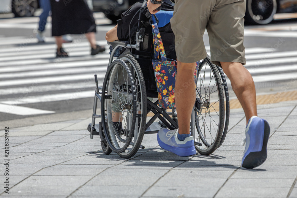 Fototapeta premium Rear view of a man pushing a wheelchair user across a city crosswalk with pedestrians and city traffic in the background.