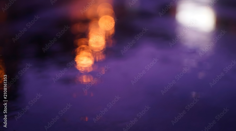 Fototapeta premium A blurred abstract image featuring warm orange bokeh lights reflecting on a dark purple wet surface, creating a dreamy and atmospheric effect.