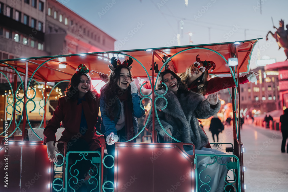 Fototapeta premium Four young women laugh together on a brightly lit festive ride at a Christmas market. The friends celebrate the holiday season on a winter evening with lights and decorations.