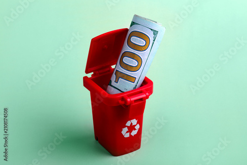 Obraz na płótnie Rolled hundred dollar bill placed inside a small red recycling bin on a mint green background, symbolizing wasteful spending or financial loss