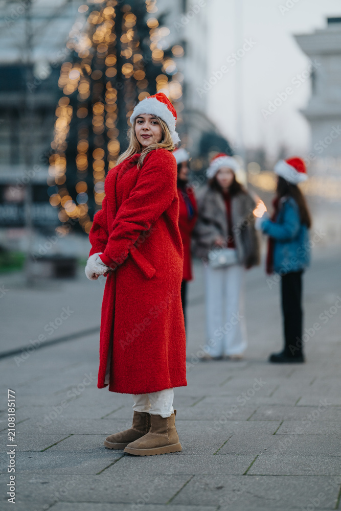 Fototapeta premium Young woman in a red coat and Santa hat stands on a city street. Three girls wearing Santa hats gather near festive holiday lights in winter.