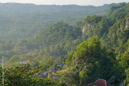 Aerial view of a lush green tropical forest valley with traditional gazebo roofs in morning mist.