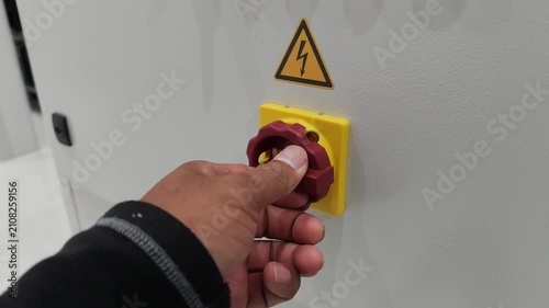 A close-up slow motion video of a technician's hand slowly turning a red and yellow main emergency switch on an electrical control panel with a high voltage warning sign