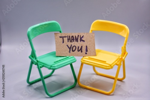 Two miniature plastic folding chairs in green and yellow hold a handwritten cardboard 