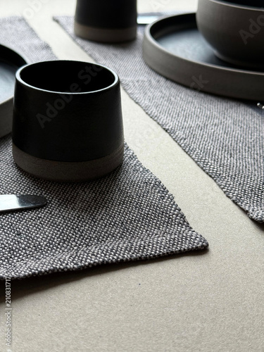 Minimal black ceramic tableware arranged on diagonally placed linen placemats. Handmade stoneware and natural textile textures create a dynamic composition for dining, home decor and interior projects