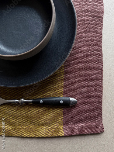 Close-up of a black ceramic bowl and plate on colorful linen with a vintage fork. Handmade stoneware and natural textile textures create a minimalist table setting for dining, home decor and interior 