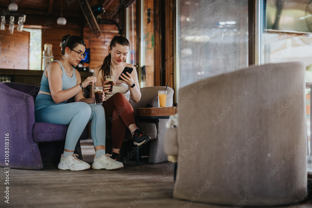 Obraz premium Two women in active wear sit in a cafe drinking iced coffee and looking at a smartphone. They relax after exercise, sharing a casual moment and a drink together.