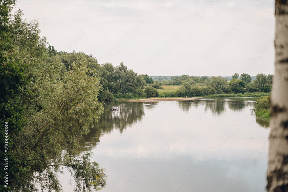 Fototapeta premium Wide scenic river with quiet current and dense wooded banks under a gloomy overcast sky, peaceful nature landscape