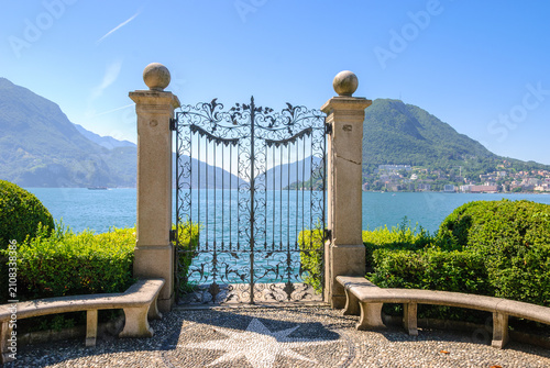 Elegant wrought iron gate overlooking Lake Lugano in Lugano, Switzerland, framed by mountains and blue sky, showcasing timeless architecture and serene lakeside scenery.