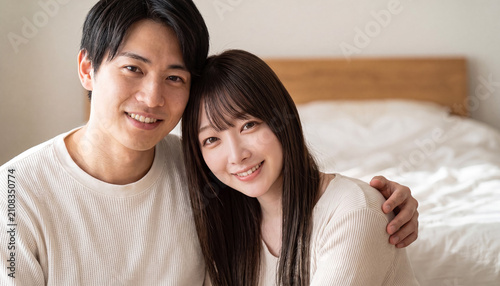Happy young Japanese couple embracing affectionately in a bright bedroom, smiling at the camera together
