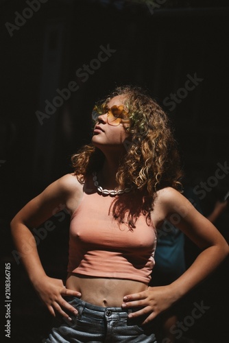 Portrait of teen girl with curly hair and heart-shaped sunglasses stands confidently outdoors, embracing the sunlight. Her hands on hips convey strength and self-assurance, creating a lively scene