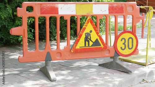 Orange construction barrier with warning sign and speed limit indicator displayed prominently on a sidewalk surrounded by greenery in an urban setting