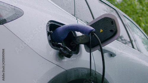 Electric vehicle charging process with a close-up view of the charging port and connector, showcasing the connection in a rainy outdoor setting with greenery in the background